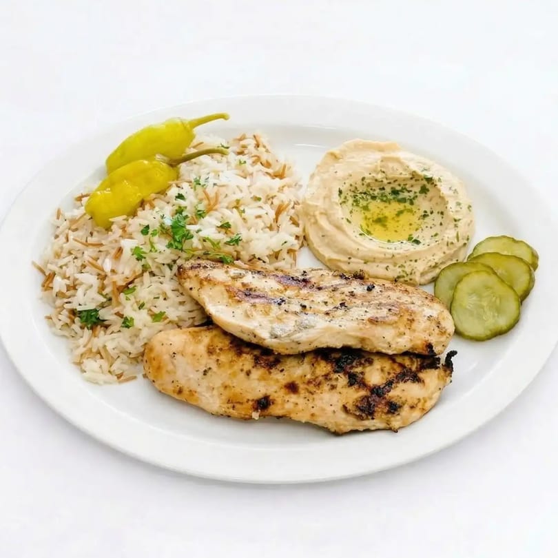 Grilled chicken with rice, hummus, pickled cucumbers, and two yellow peppers on a white plate.
