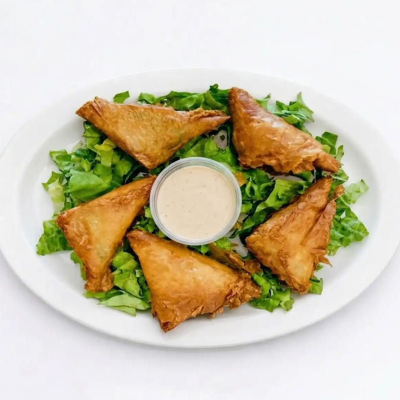 Five golden-brown triangular pastries, possibly samosas or spanakopita, arranged on a bed of fresh green lettuce with a small cup of dipping sauce in the center.